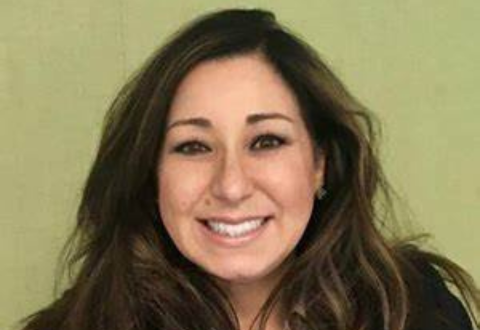 Woman with long brunette hair smiling in front of a green background. She is wearing a black cardigan