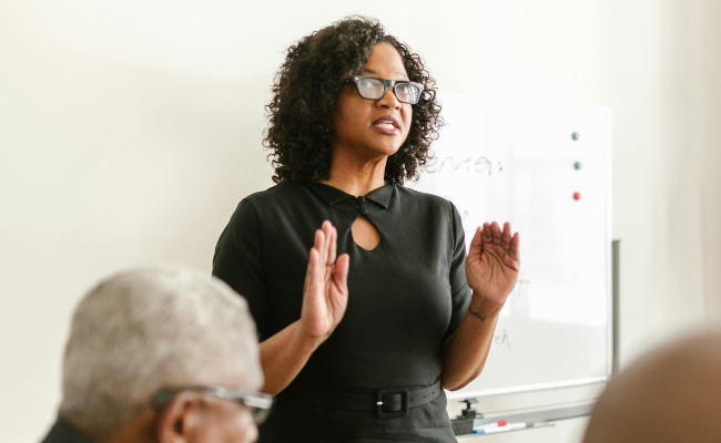 Woman in business attire stands speaking in front of a seated group of people